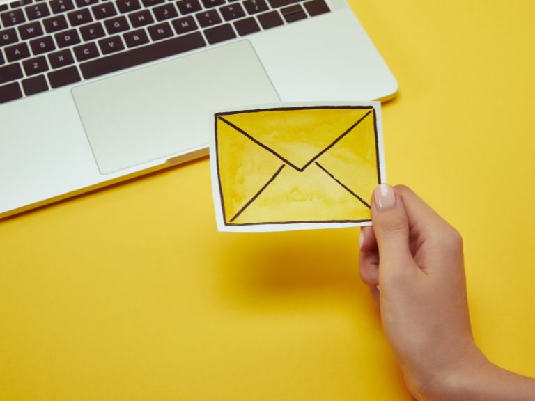 Hand holding illustration of an envelope with a laptop behind it, against a yellow background